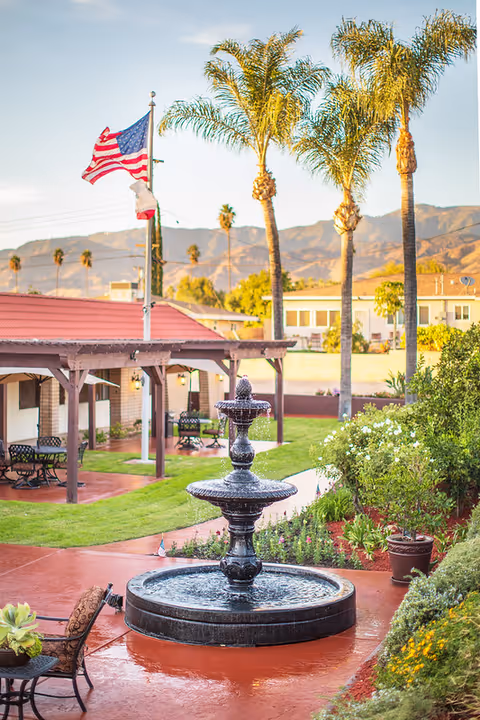 Outdoor courtyard featuring a tiered fountain, palm trees, an American flag, and covered seating.