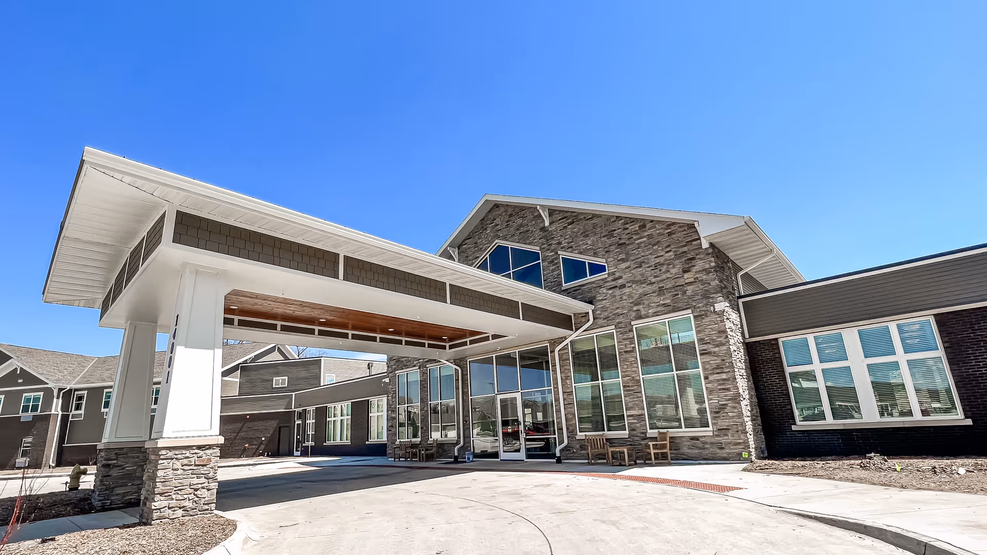 Exterior view of Edencrest at Timberline senior living facility showing the main entrance with a covered drop-off area, stone and brick facade, large windows, and clear blue sky.
