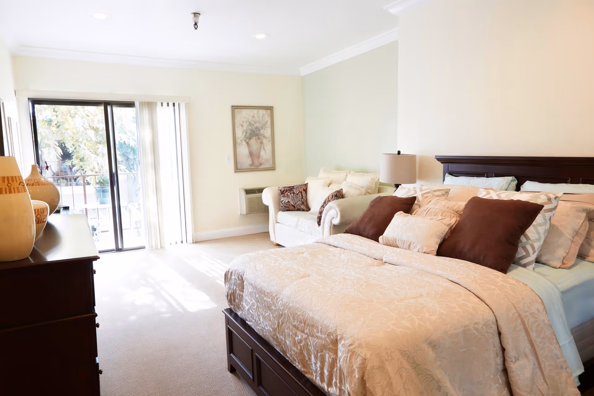 Bright bedroom with a large bed covered in beige and brown pillows and a matching comforter. There is a white loveseat with decorative pillows against the wall, a wooden dresser with vases on top, a lamp on a bedside table, and a sliding glass door leading to a balcony with trees visible outside.