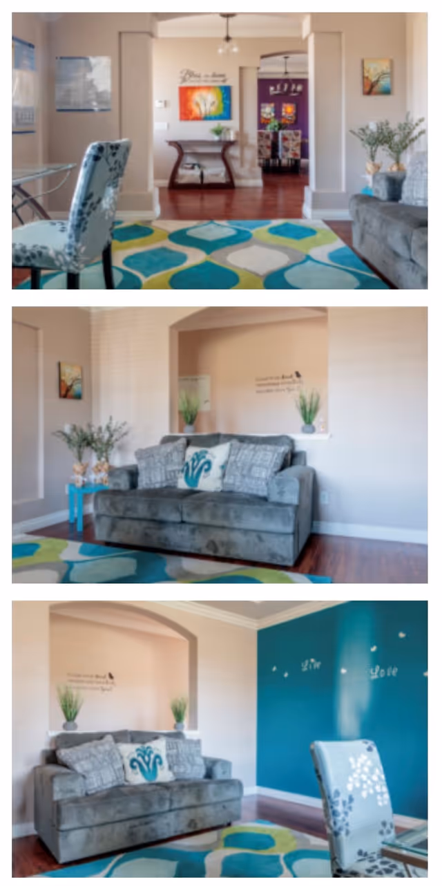 A cozy living room area in a senior living facility featuring a gray sofa with patterned cushions, a colorful geometric area rug in shades of blue, green, and white, and a glass-top table with a floral-patterned chair. The walls are decorated with framed artwork, plants, and inspirational wall decals. The room has wooden flooring and an open layout leading to a dining area with floral upholstered chairs.
