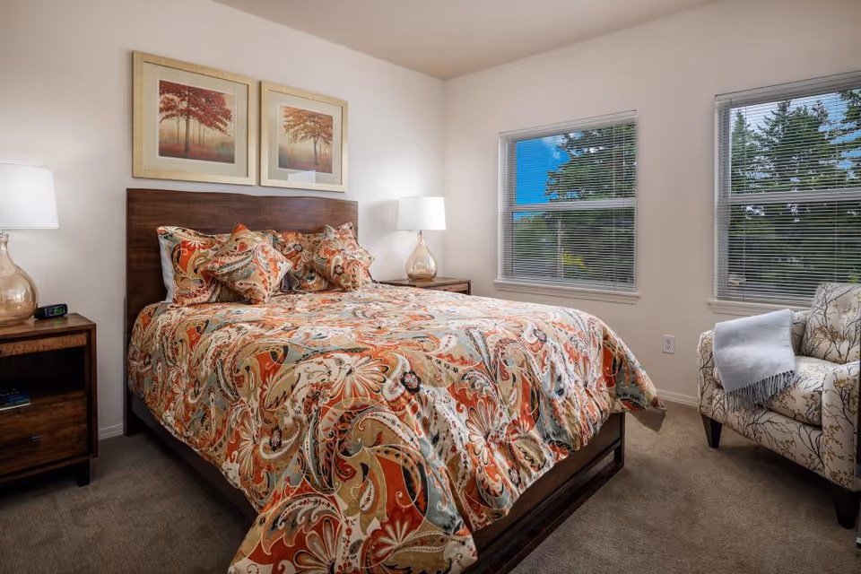 A cozy bedroom with a large bed covered in a colorful paisley-patterned bedspread and matching pillows. The room has two wooden nightstands with glass lamps on each side of the bed. Two framed pictures of trees hang above the headboard. There are two windows with blinds letting in natural light, and a patterned armchair with a folded blanket draped over one arm is positioned near the windows.