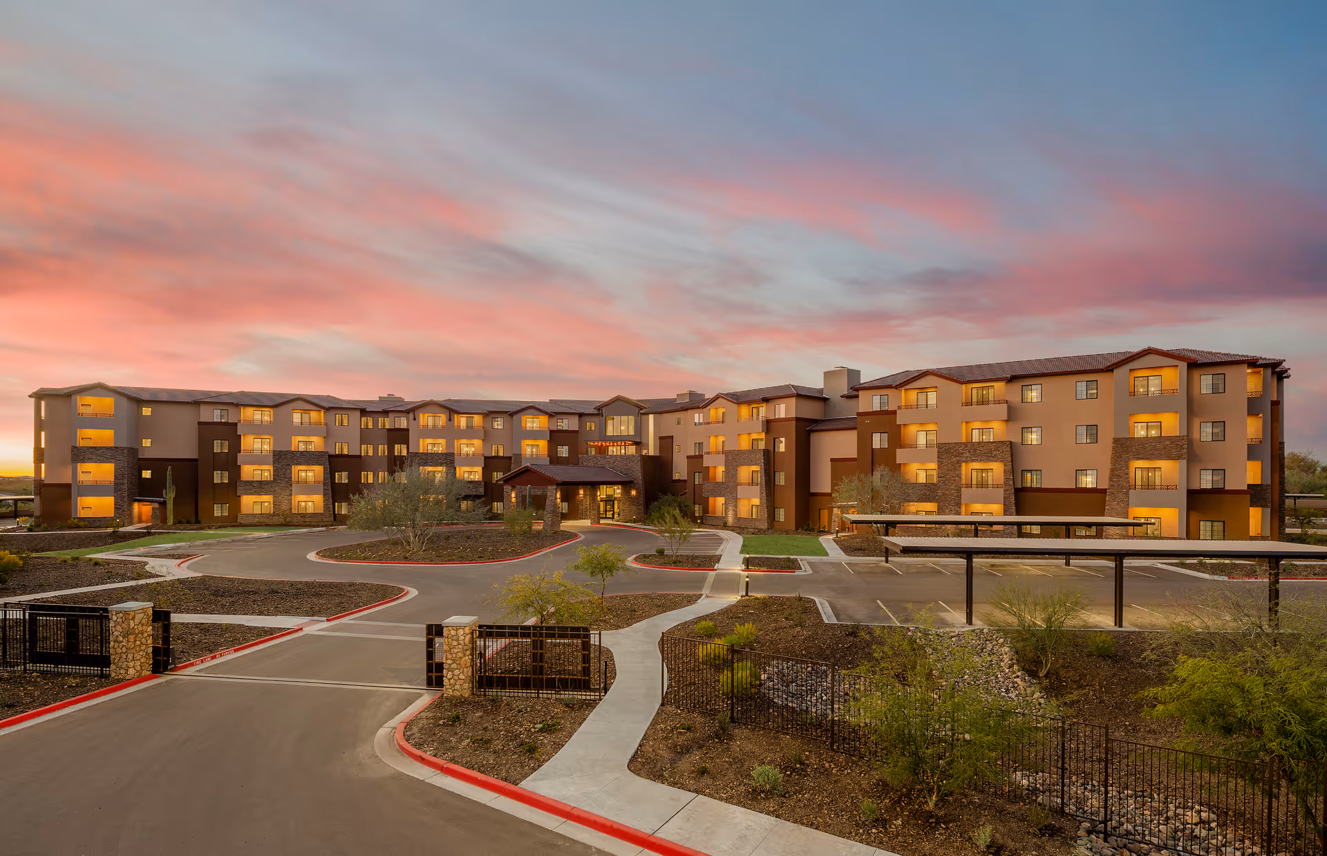 Wide exterior view of a multi-story senior living building at dusk with lit windows and a landscaped circular driveway.