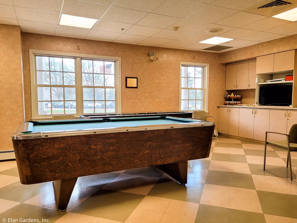 A pool table in a community recreation room with windows, cabinets, chairs, and a TV.