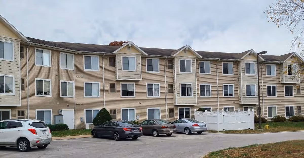 Exterior view of a three-story beige apartment building with multiple windows and a parking lot in front containing several parked cars. The sky is cloudy and there is some greenery around the building.