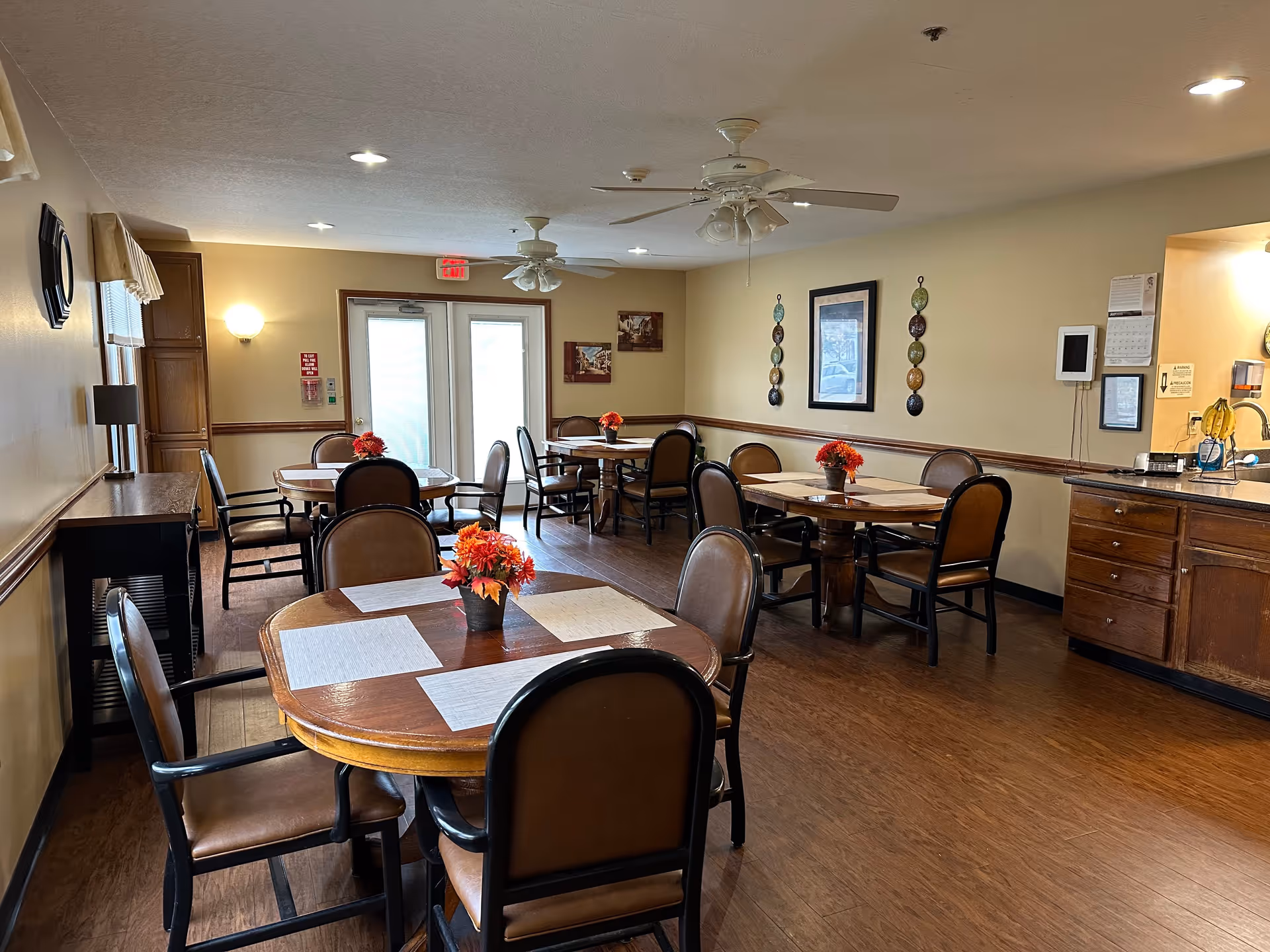 A dining room with several round wooden tables, each surrounded by brown cushioned chairs. Each table has a small flower arrangement with red and orange flowers. The room has wooden flooring, beige walls with framed pictures and decorative wall hangings, ceiling fans with lights, and a counter with cabinets on the right side. There is a double door with frosted glass at the far end of the room.