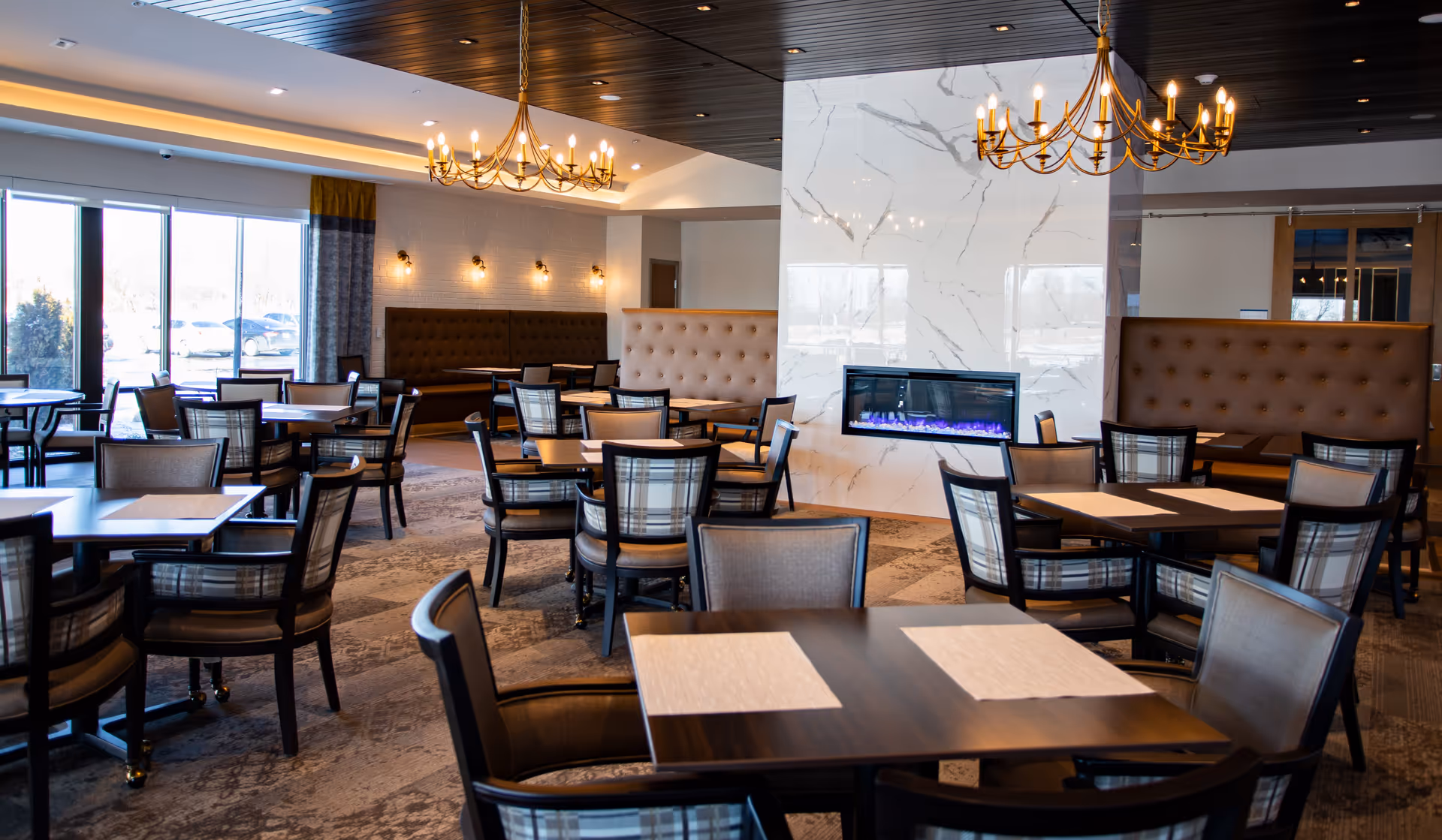 A spacious dining room with multiple tables and chairs arranged neatly. The room features large windows allowing natural light to enter, two elegant chandeliers hanging from a dark wooden ceiling, and a modern electric fireplace set in a white marble wall. Upholstered bench seating lines the walls, and the overall decor is warm and inviting.