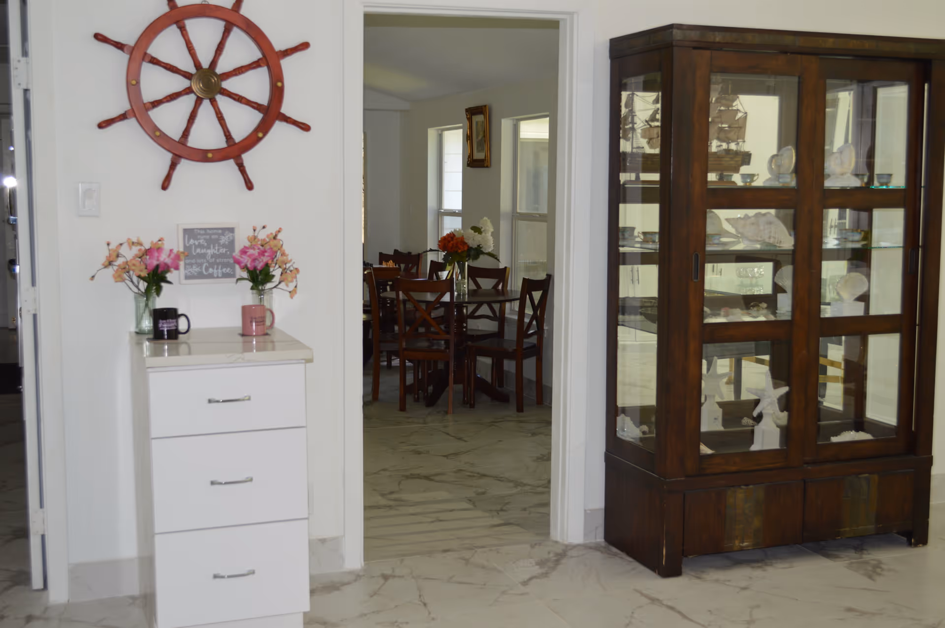 Interior view showing a small white cabinet with flowers and a ship's wheel on the wall, a glass display cabinet, and a dining table with chairs visible through a doorway.
