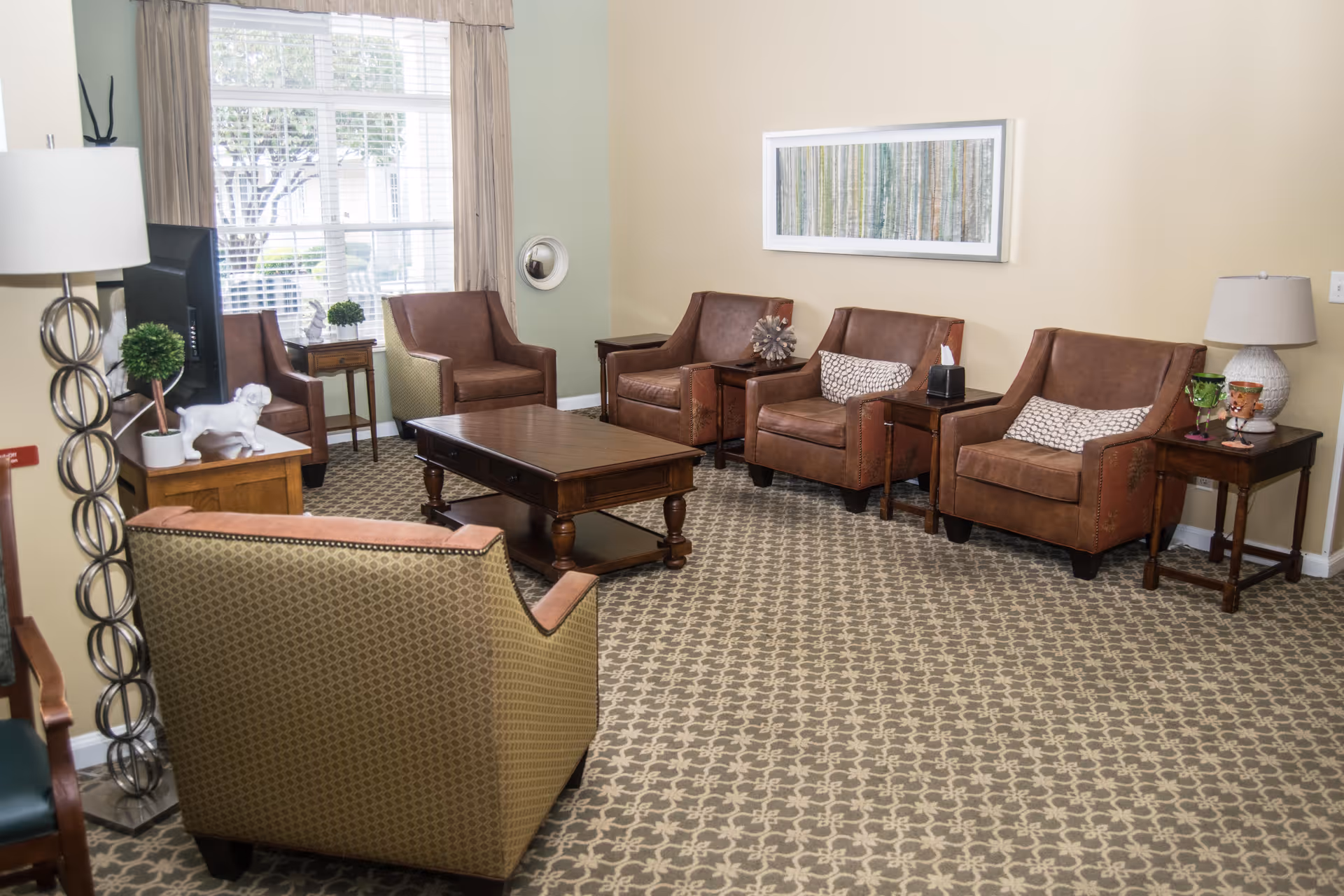 A cozy senior living community lounge with a patterned carpet, four brown leather armchairs with pillows, a wooden coffee table, side tables with lamps and decorative items, a large window with beige curtains, and a framed artwork on the wall.