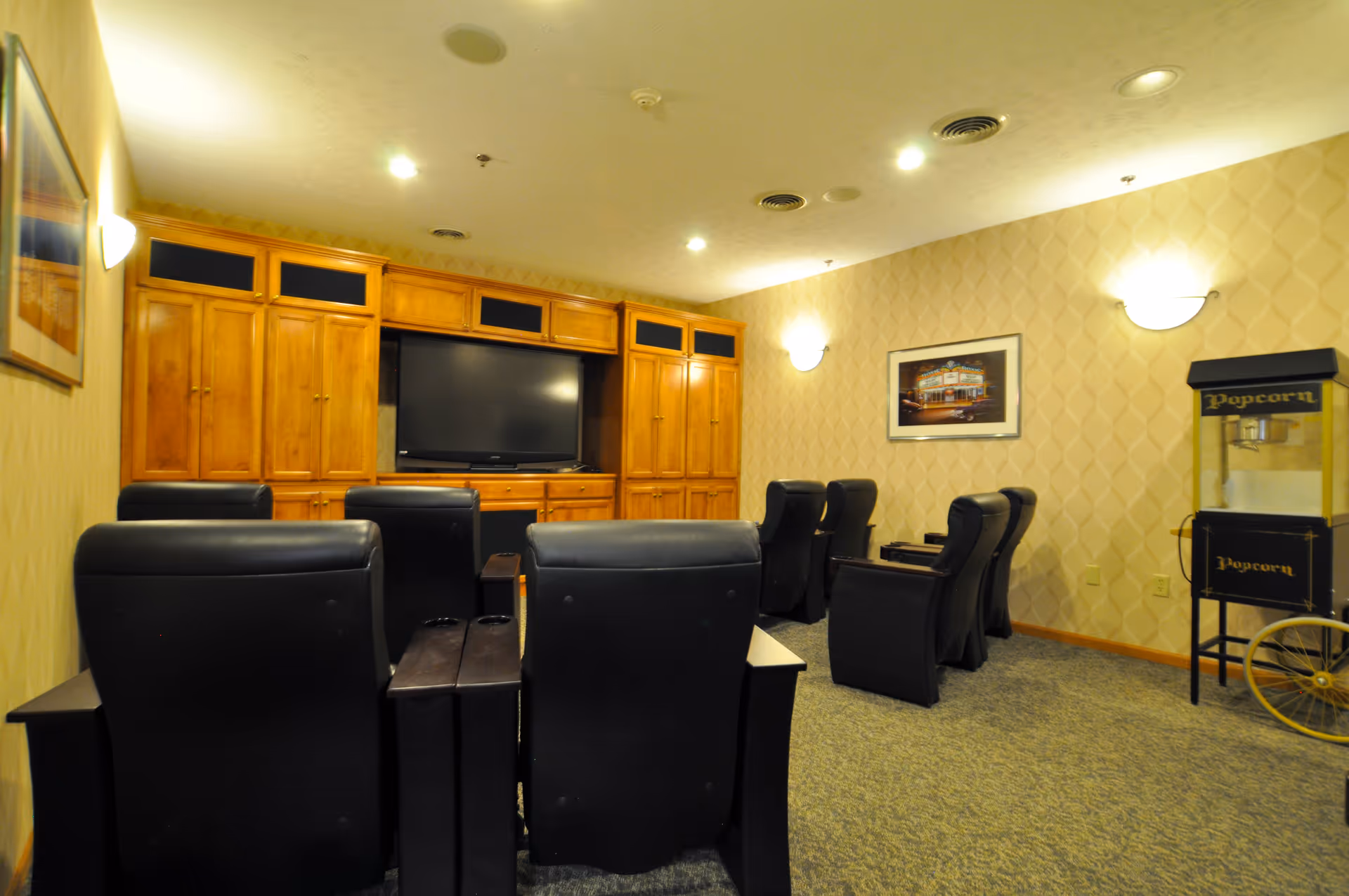 A cozy media room with several black leather recliner chairs arranged in rows facing a large flat-screen TV mounted on a wooden entertainment center. The room has beige patterned wallpaper, carpeted floor, wall-mounted lights, a framed picture on the wall, and a vintage-style popcorn machine in the corner.