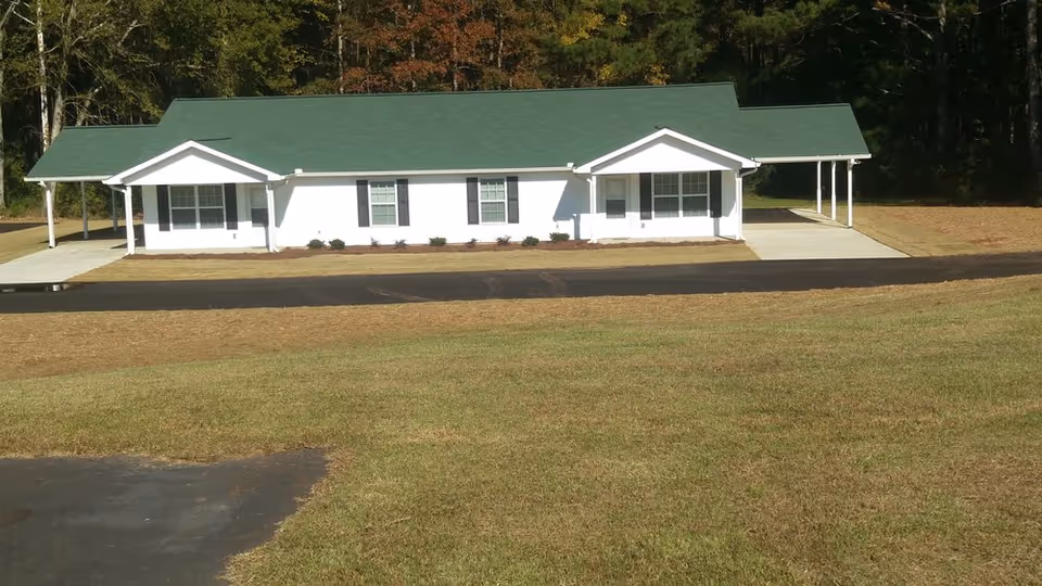 Single-story white building with a green roof, two covered carports, and a grassy lawn in front.