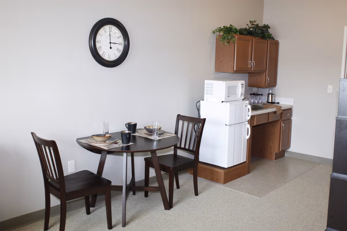 A small dining area with a round dark wooden table set for two with bowls, mugs, and glasses. Two matching wooden chairs are placed around the table. On the wall above the table is a large round clock with Roman numerals. To the right is a compact kitchenette with wooden cabinets, a white mini refrigerator, a microwave on top of the refrigerator, a sink, and some glassware. There are green plants on top of the cabinets.