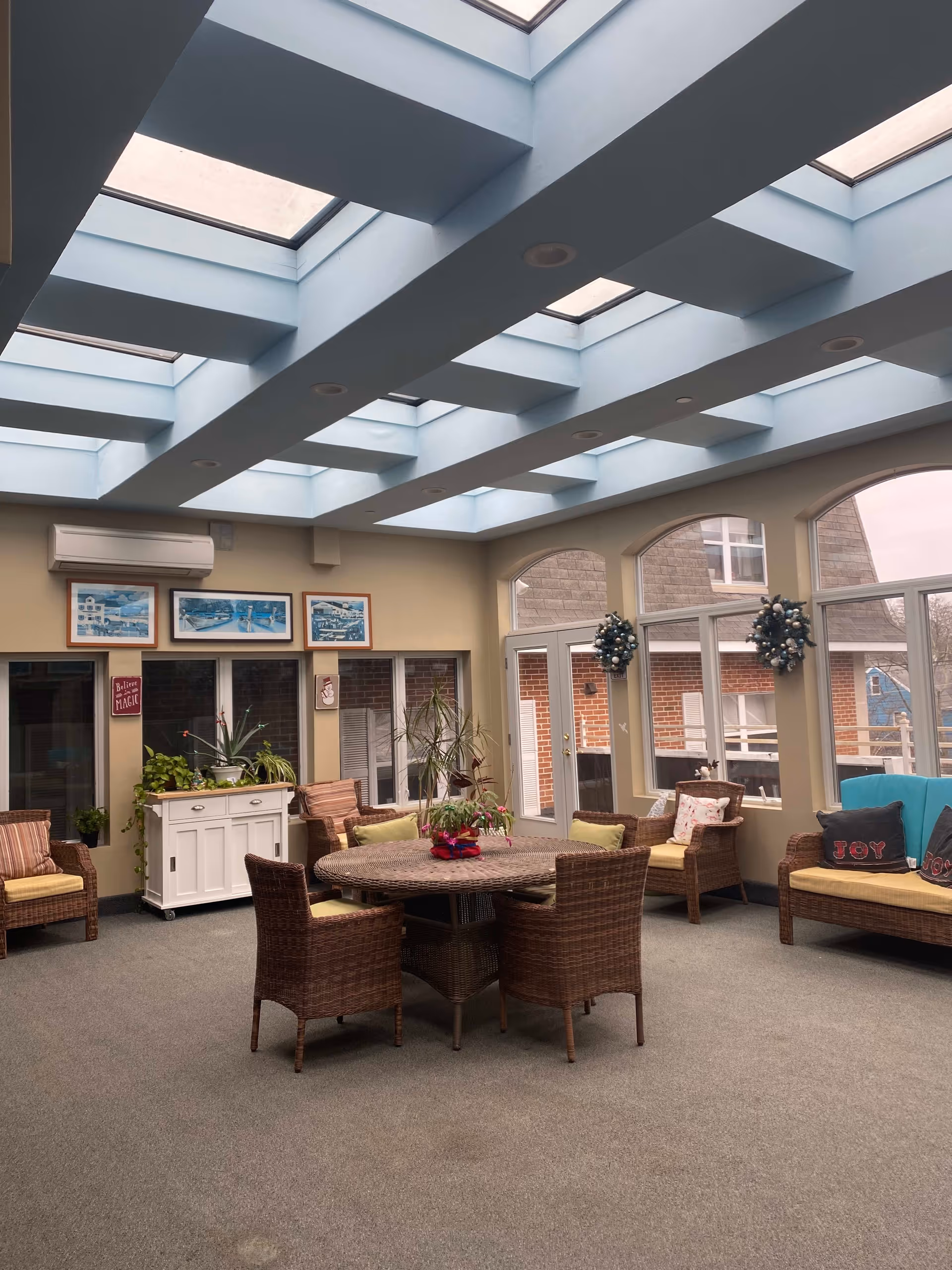 A bright and spacious common area with a high ceiling featuring multiple skylights. The room has wicker chairs and a round wicker table in the center, along with additional seating along the walls. There are large windows and glass doors letting in natural light, decorated with holiday wreaths. The walls have framed pictures and a small white cabinet with plants on top.
