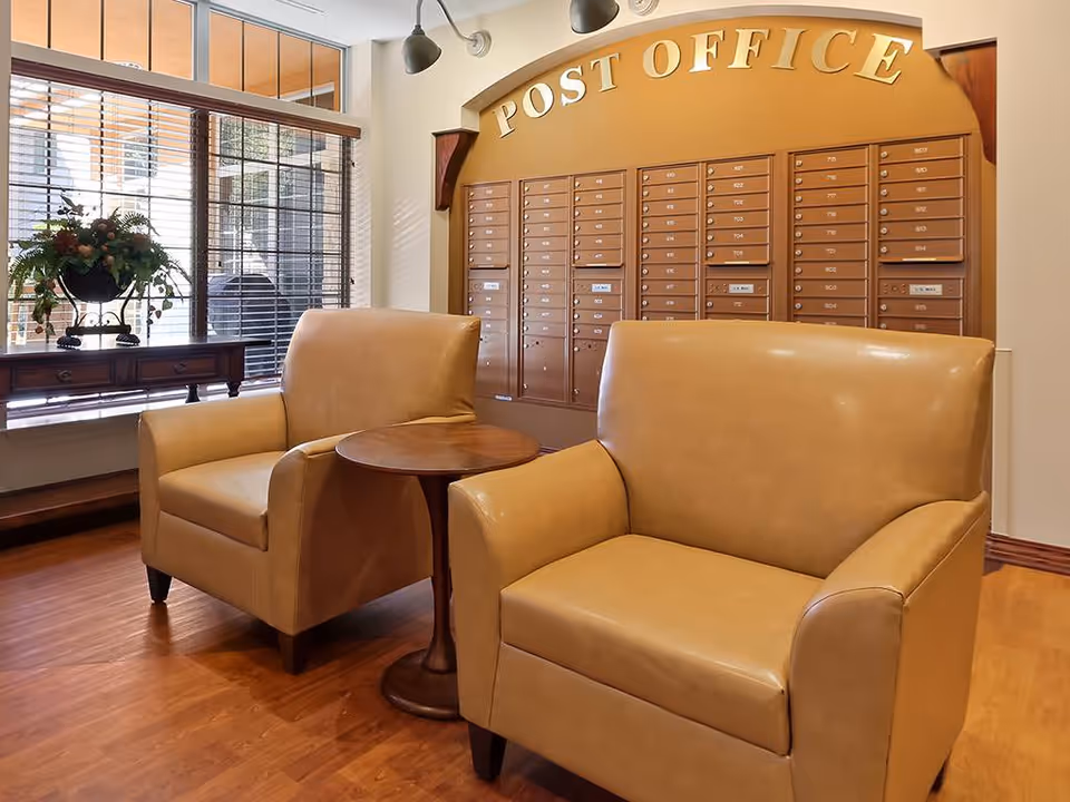 Interior view of a post office area inside a facility with two tan leather armchairs and a small round wooden table between them. Behind the chairs is a wall with multiple mailboxes labeled with numbers, and above the mailboxes is a sign that reads 'POST OFFICE'. To the left, there is a window with blinds and a table with a decorative plant on it.