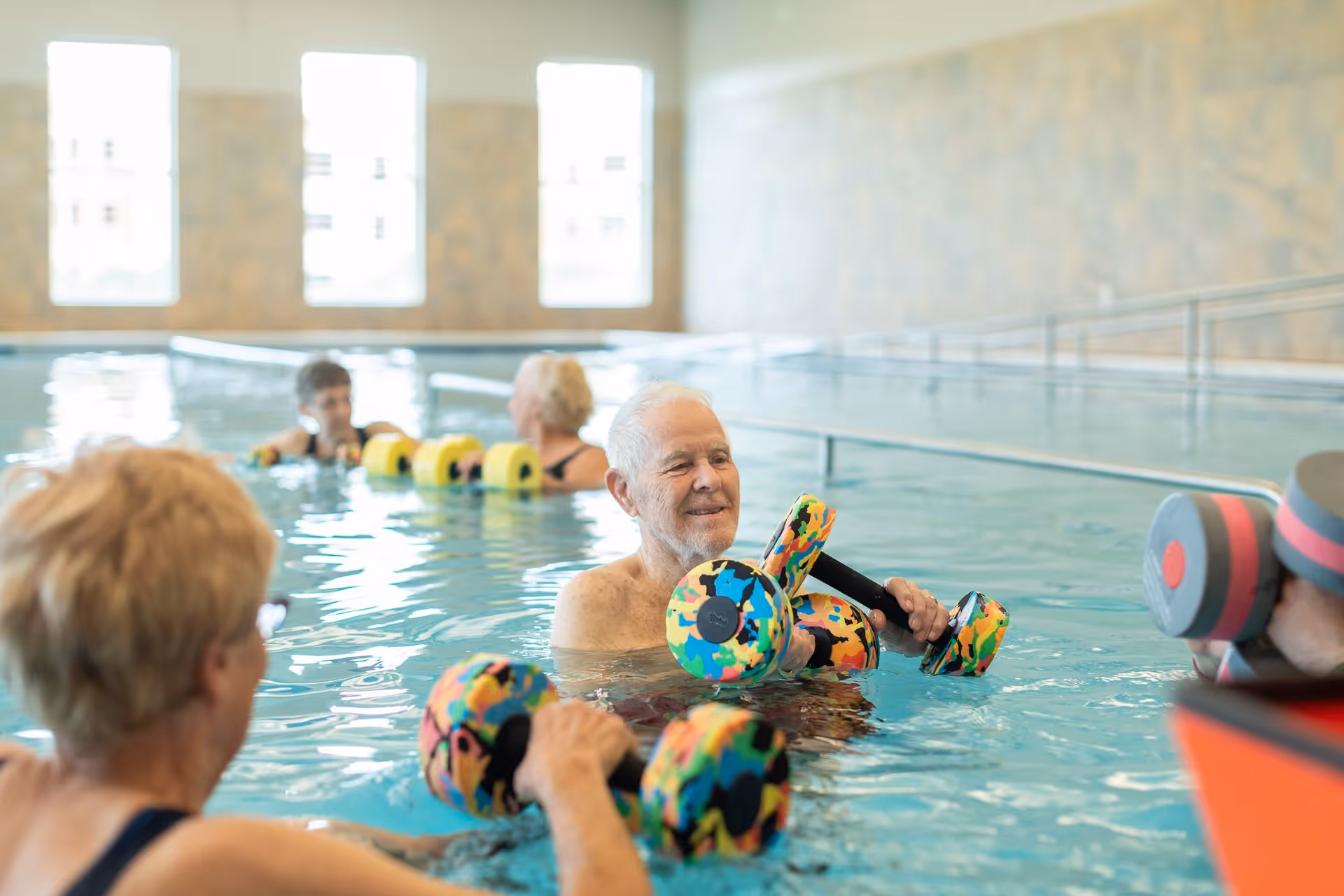 A group of elderly people participating in a water exercise class in an indoor swimming pool, using colorful foam dumbbells for resistance training.