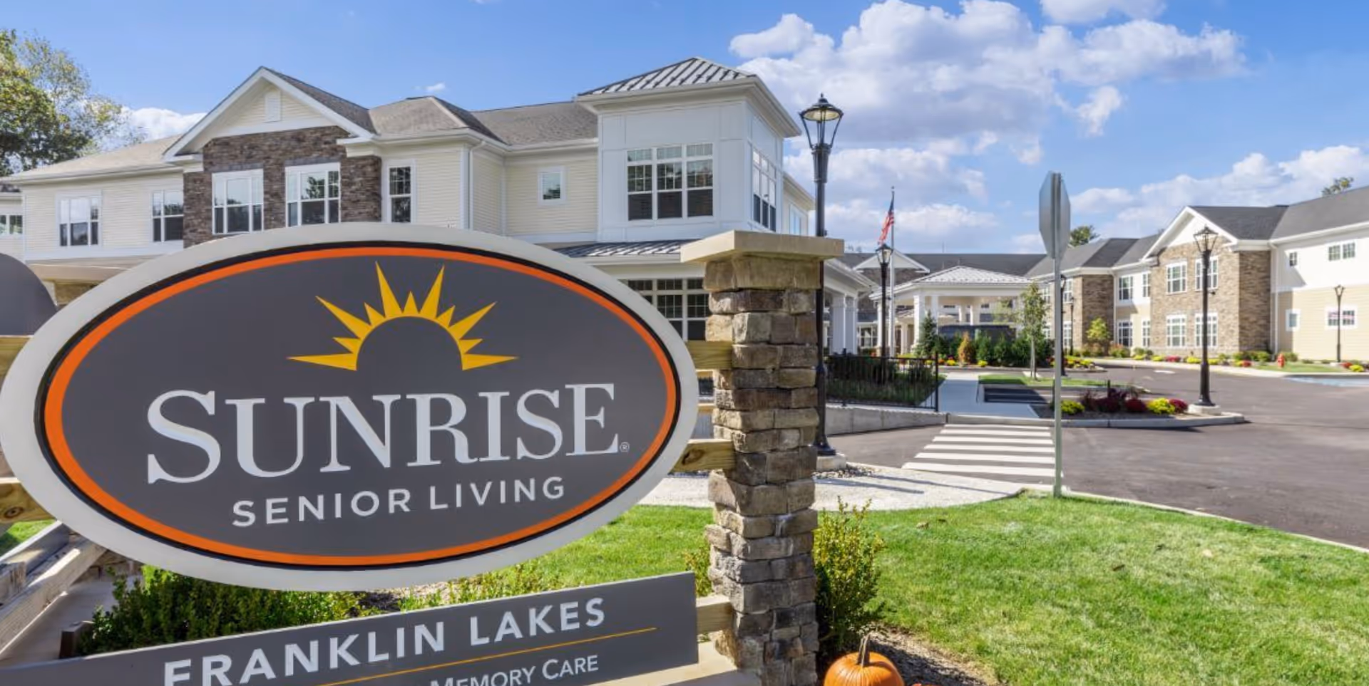 Exterior view of Sunrise Senior Living facility in Franklin Lakes, showing the main building, a large sign with the Sunrise logo, a well-maintained lawn, and a clear blue sky with some clouds.