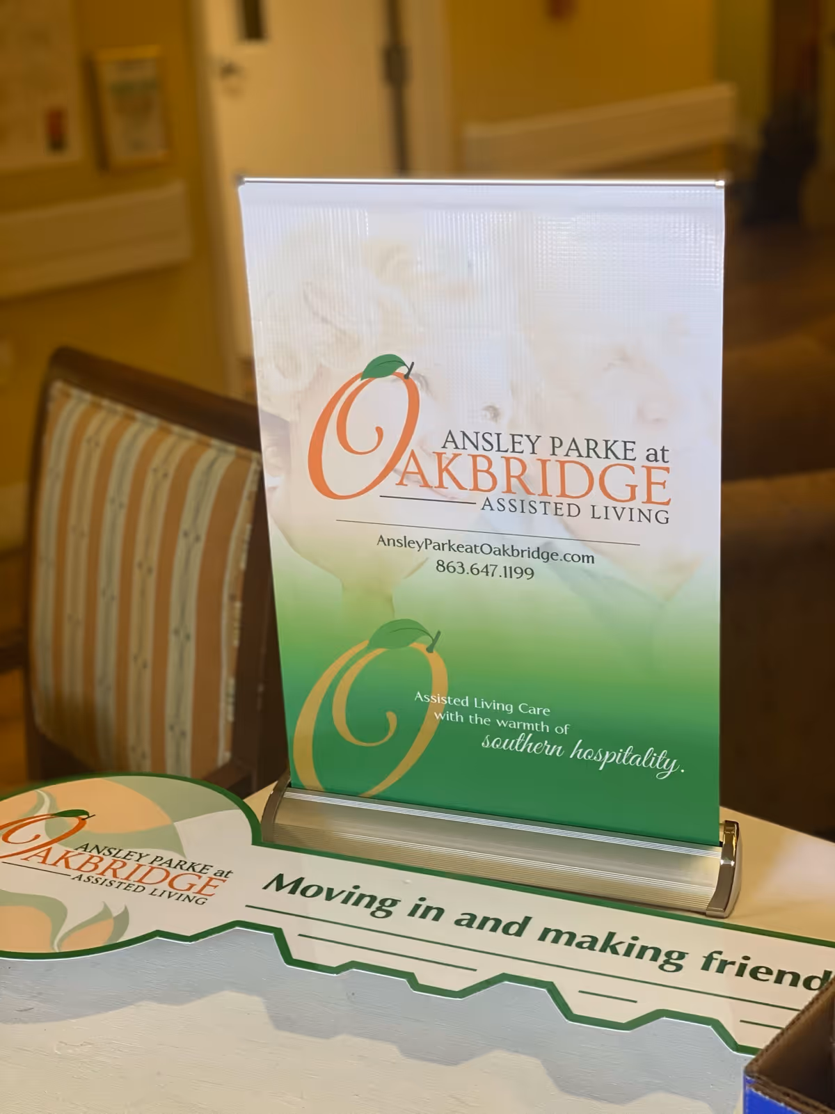 A table display at Ansley Parke at Oakbridge Assisted Living featuring a standing sign with the facility's logo, website, phone number, and the slogan 'Assisted Living Care with the warmth of southern hospitality.' A decorative key-shaped sign on the table reads 'Moving in and making friends.'