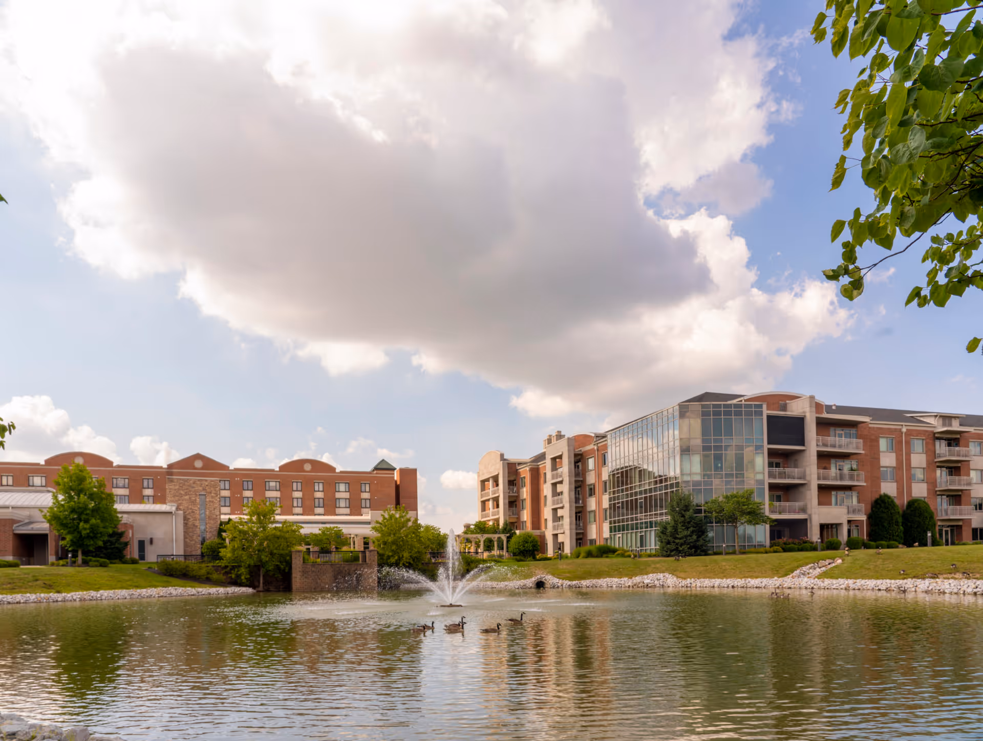 Exterior view of a senior living campus with brick buildings, a glass atrium, and a pond.
