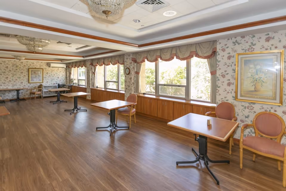 A spacious dining room with wooden floors, several square wooden tables with black metal legs, and pink cushioned chairs. The room has large windows with floral curtains allowing natural light to enter. The walls are decorated with floral wallpaper and framed artwork. The ceiling features recessed lighting and chandeliers.