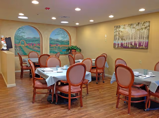 Dining room with several tables covered with light blue tablecloths, each set with cups, saucers, and utensils. The room has wooden chairs with cushioned backs and seats. The walls are decorated with a mural of an outdoor garden scene and a framed painting of birch trees. The floor is wood, and the ceiling has recessed lighting.