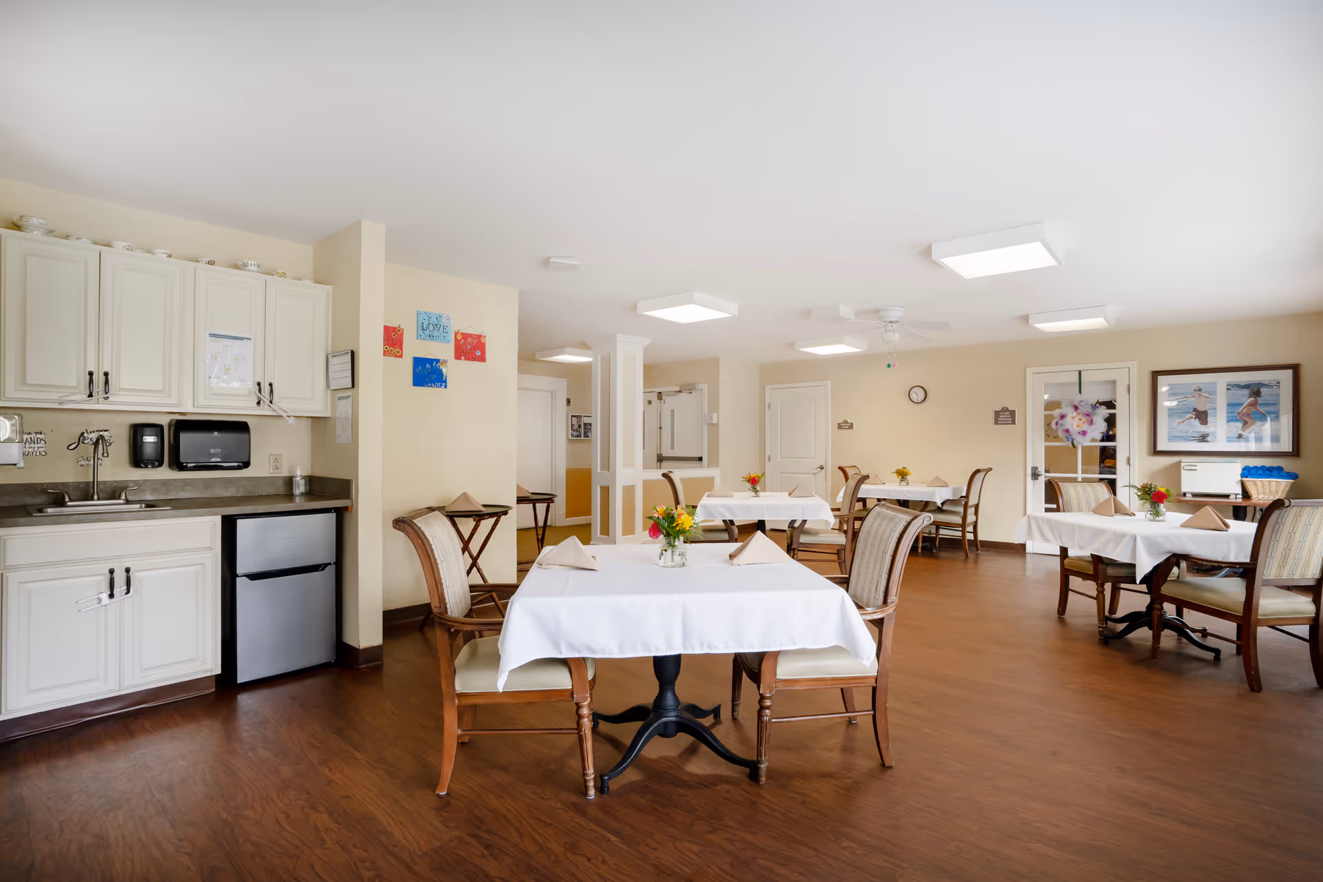 A bright dining room with several tables covered in white tablecloths, each set with folded napkins and small flower arrangements. The room has wooden flooring, beige walls, and ceiling lights. There is a small kitchenette area with white cabinets, a sink, and a mini refrigerator on the left side. The room appears clean and inviting with framed pictures on the walls and a clock above a door in the background.