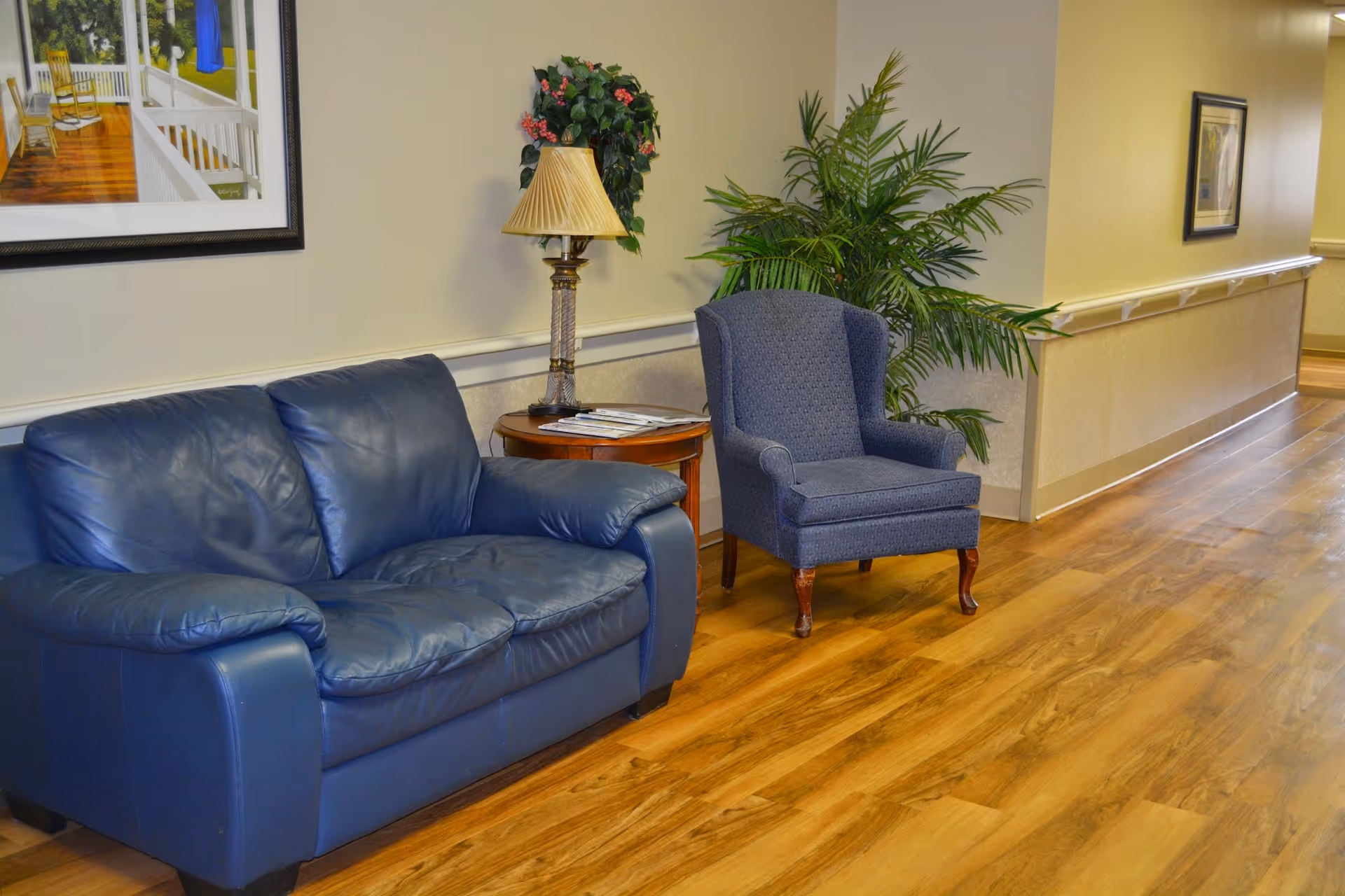 A cozy seating area in a hallway with a blue leather couch, a blue upholstered armchair, a wooden side table with a lamp and magazines, a potted plant, and framed artwork on the walls. The floor is wooden and the walls are painted beige.