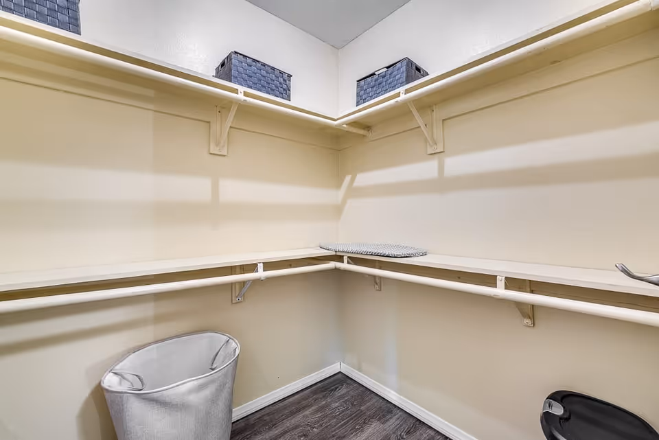 Empty walk-in closet with white shelves and hanging rods along the walls, two gray storage baskets on the upper shelf, a gray laundry hamper on the floor, and an ironing board resting on the shelf.