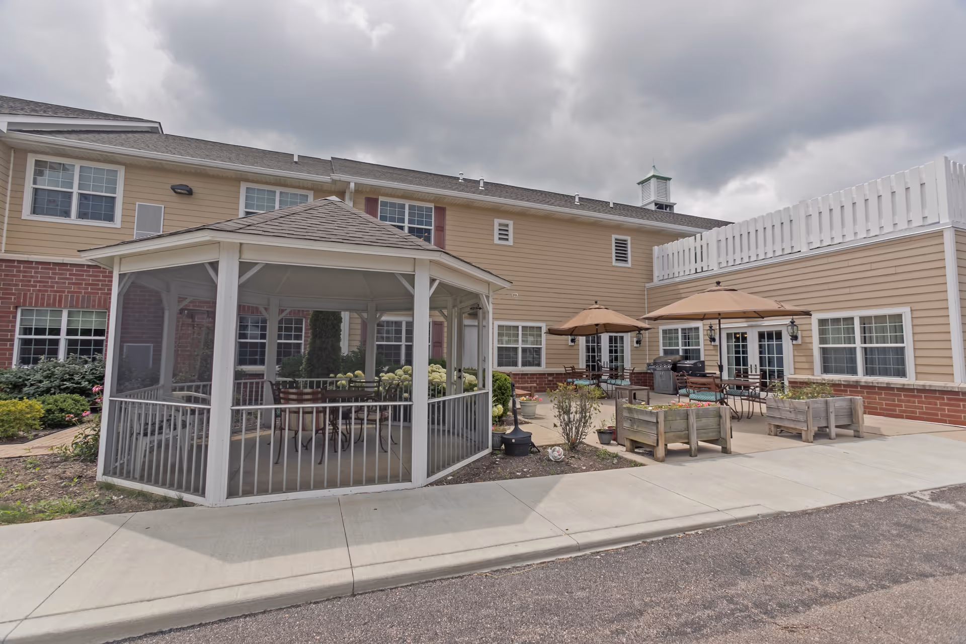 Outdoor patio area of a senior living facility with a white gazebo, several tables with umbrellas, chairs, and planter boxes. The building exterior is beige with red brick accents and multiple windows under a cloudy sky.