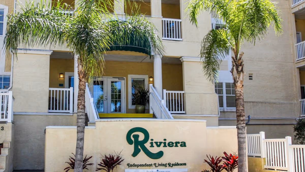 Exterior view of the entrance to Riviera Senior Living, an independent living residence. The building is light yellow with white railings and columns. There are two palm trees in front of the entrance and a sign on the wall that reads 'Riviera Independent Living Residences'.