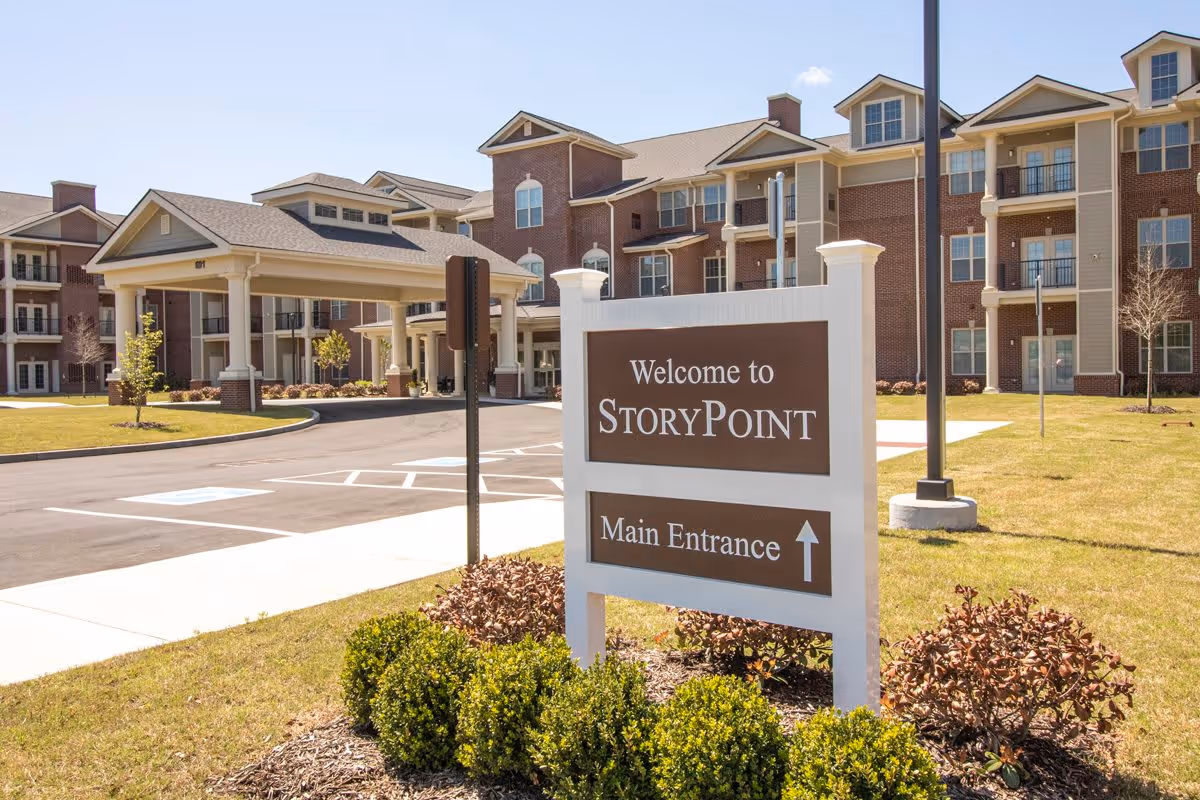 Exterior view of StoryPoint Collierville senior living facility showing the main entrance with a covered driveway, a large brick building with multiple balconies, and a sign that reads 'Welcome to StoryPoint Main Entrance' surrounded by landscaping and a clear sky.