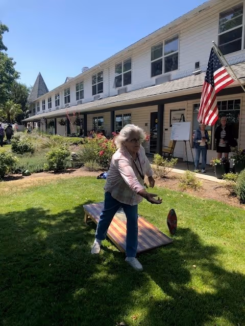 An elderly woman playing cornhole on a grassy lawn outside a two-story assisted living facility building. The building has white siding, multiple windows, and an American flag displayed near the entrance. Several people are standing near the building's entrance in the background.