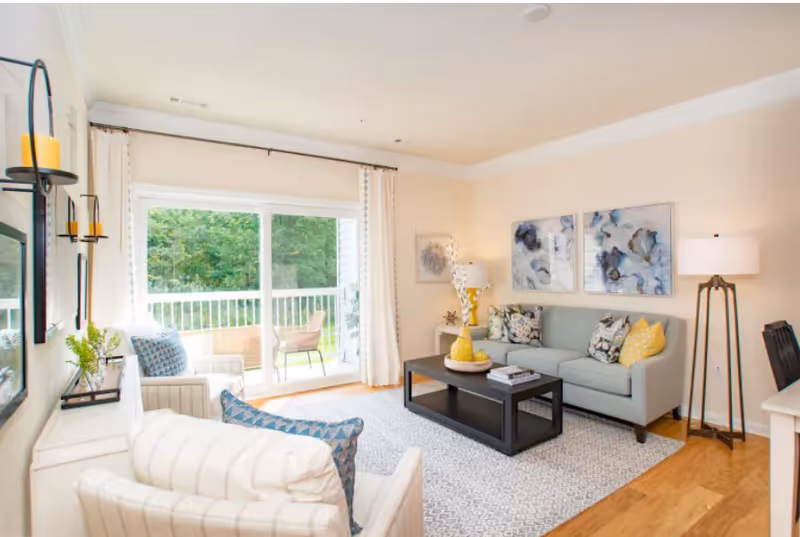 Bright living room with light-colored walls and wooden floor. The room features a light gray sofa with patterned and yellow pillows, a black coffee table with decorative items, two armchairs with blue patterned pillows, a floor lamp, and wall art. Large sliding glass doors open to a balcony with outdoor chairs and a view of green trees.