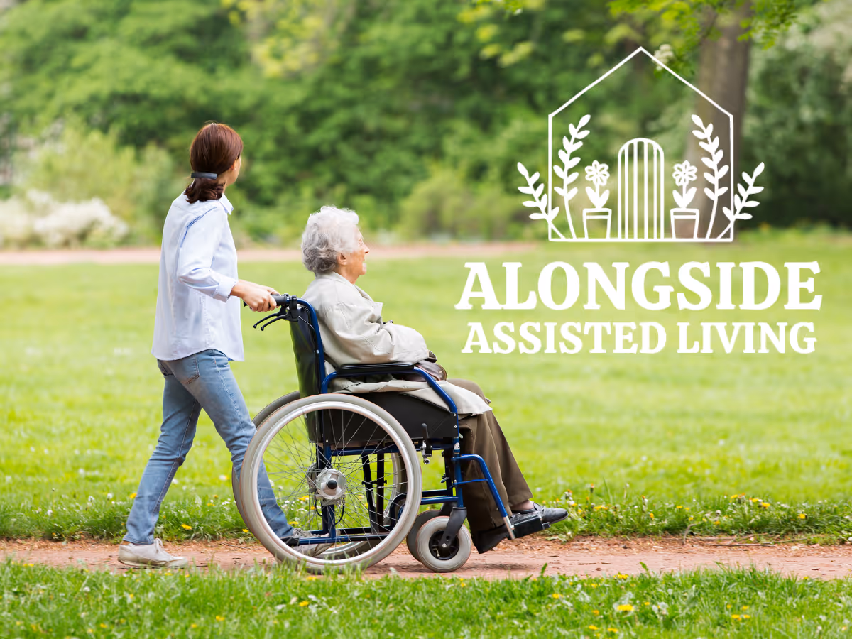 A caregiver pushing an elderly woman in a wheelchair along a path in a green park with trees in the background. The logo for Alongside Assisted Living is overlaid on the right side of the image.