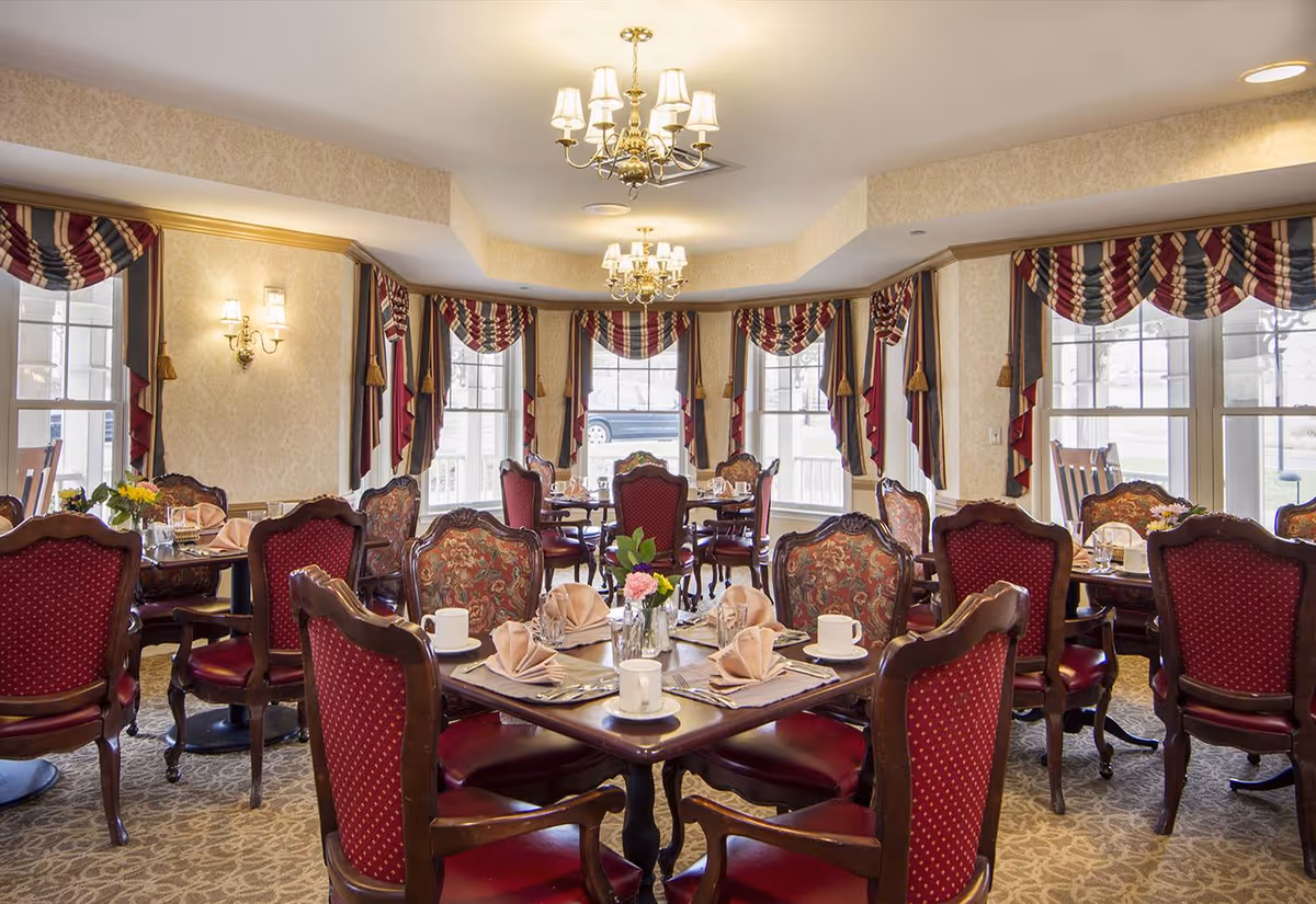 A dining room with multiple wooden tables set with beige napkins, white cups, and small flower arrangements. The chairs have red upholstery with wooden frames, and the room features large windows with red, blue, and beige draped curtains. The walls are light-colored with sconces and chandeliers providing warm lighting.