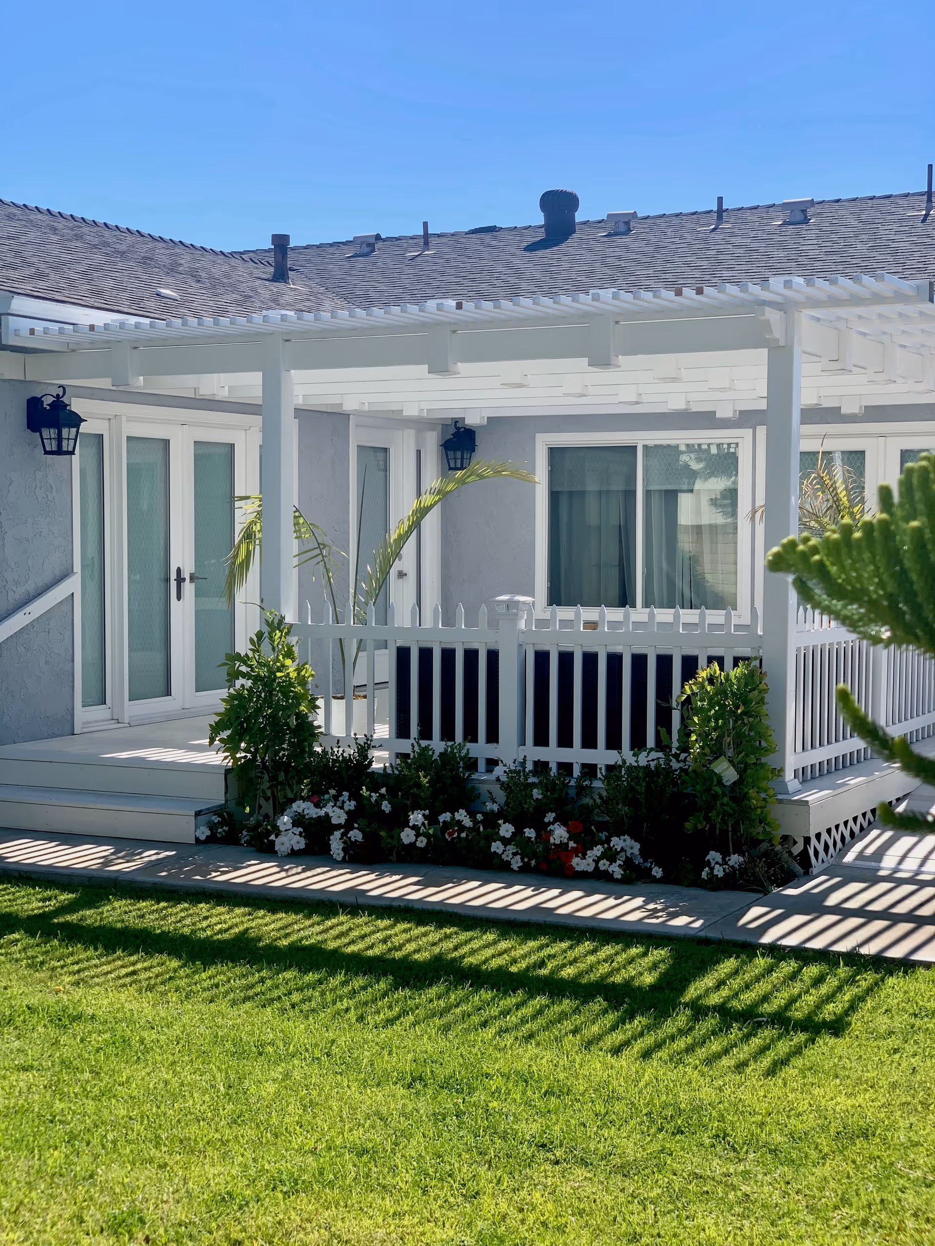 A bright outdoor patio area with a white pergola attached to a light gray building. The patio is surrounded by a white picket fence and has a small garden bed with green shrubs and white and red flowers. The lawn in front is green and well-maintained under a clear blue sky.