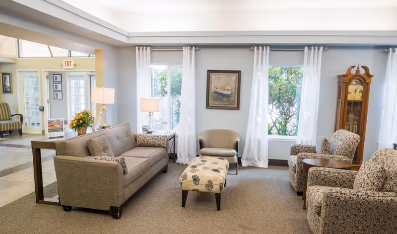 A bright and cozy living room area with a gray sofa, patterned armchairs, a small upholstered ottoman, and a wooden grandfather clock. Large windows with sheer white curtains let in natural light, and a framed painting of a boat hangs on the wall between the windows. There are two table lamps on side tables next to the sofa, and a small table with flowers is visible in the adjacent room.