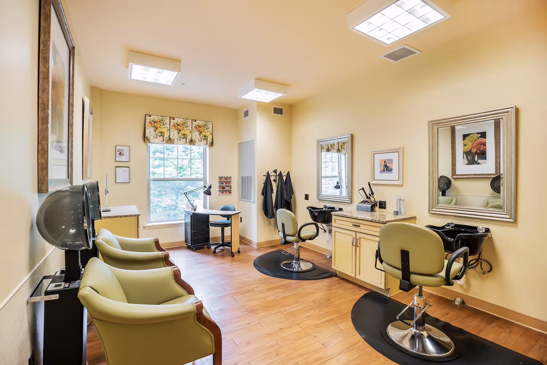 Bright salon room with styling chairs, shampoo bowls, hair dryers, mirrors, and a desk by a window.