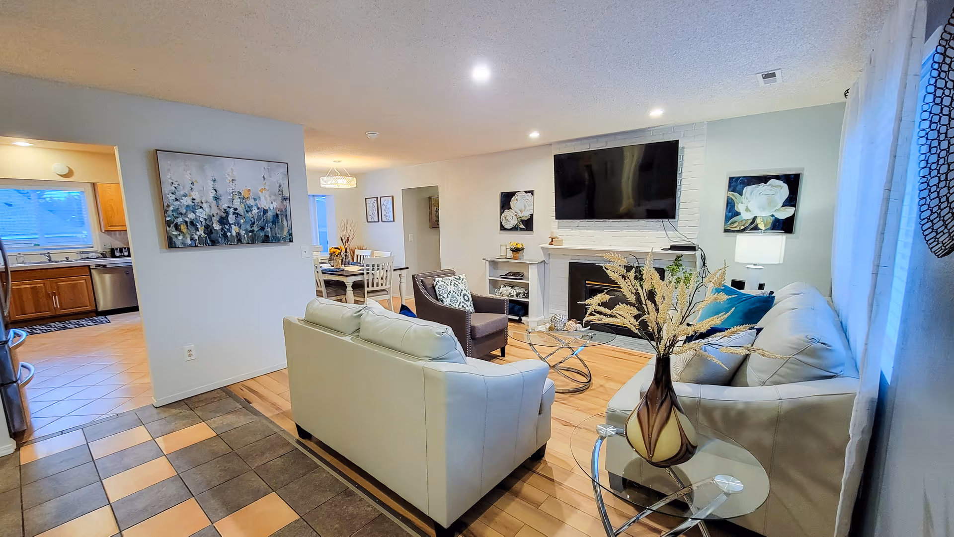 A cozy living room with two light-colored sofas, a glass coffee table, a fireplace with a mounted flat-screen TV above it, and decorative artwork on the walls. The room has wooden flooring and is adjacent to a dining area with a table and chairs, and a kitchen with wooden cabinets and tiled floor is visible in the background.