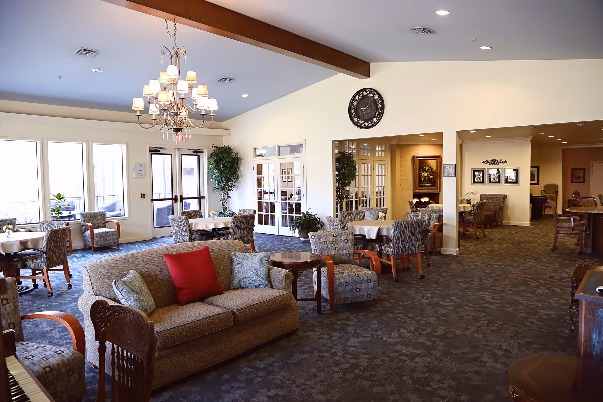 A spacious and well-lit common area in a retirement community featuring a beige sofa with colorful pillows, several armchairs, round tables with chairs, large windows, a chandelier, and decorative plants. The room has a carpeted floor and light-colored walls with framed artwork and a large clock.