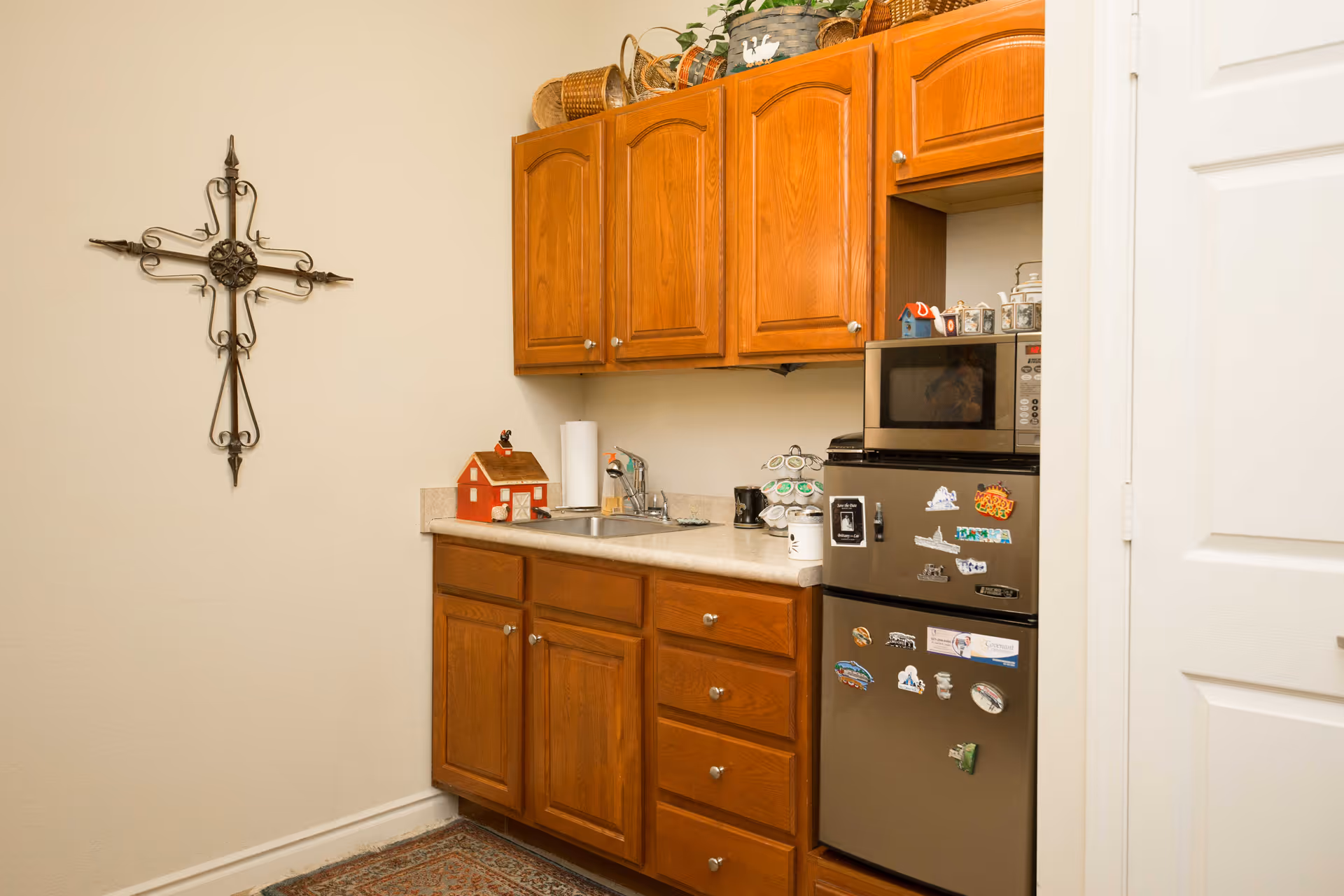 A small kitchen area with wooden cabinets, a countertop with a sink, a mini refrigerator covered with magnets, and a microwave on top of the refrigerator. There is a decorative metal cross hanging on the wall to the left and some baskets and plants on top of the cabinets.