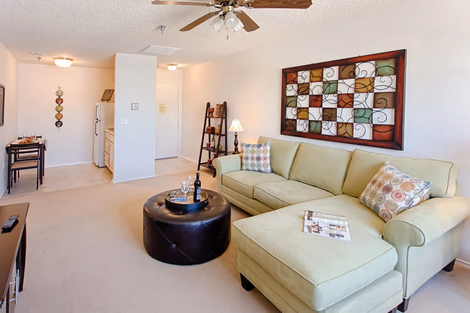 Bright living room with a light-green sectional sofa, round ottoman coffee table, decorative wall art, and a small kitchenette and dining area in the background.