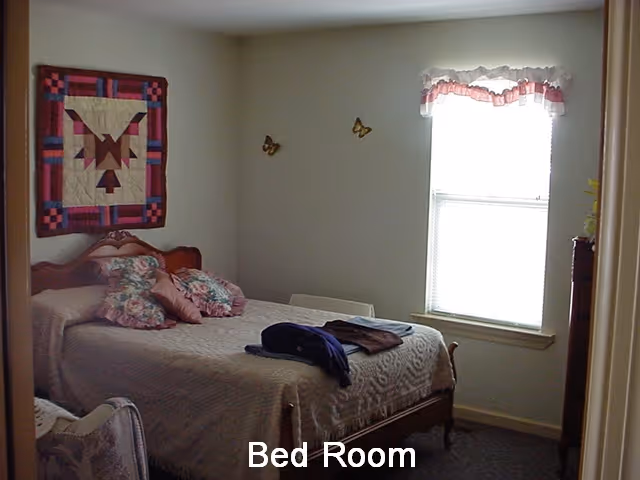 A bedroom with a bed covered in a beige quilt and several floral pillows. There is a colorful quilt hanging on the wall above the bed and two butterfly decorations on the adjacent wall. A window with a white and pink valance lets in natural light. A chair is partially visible in the foreground.