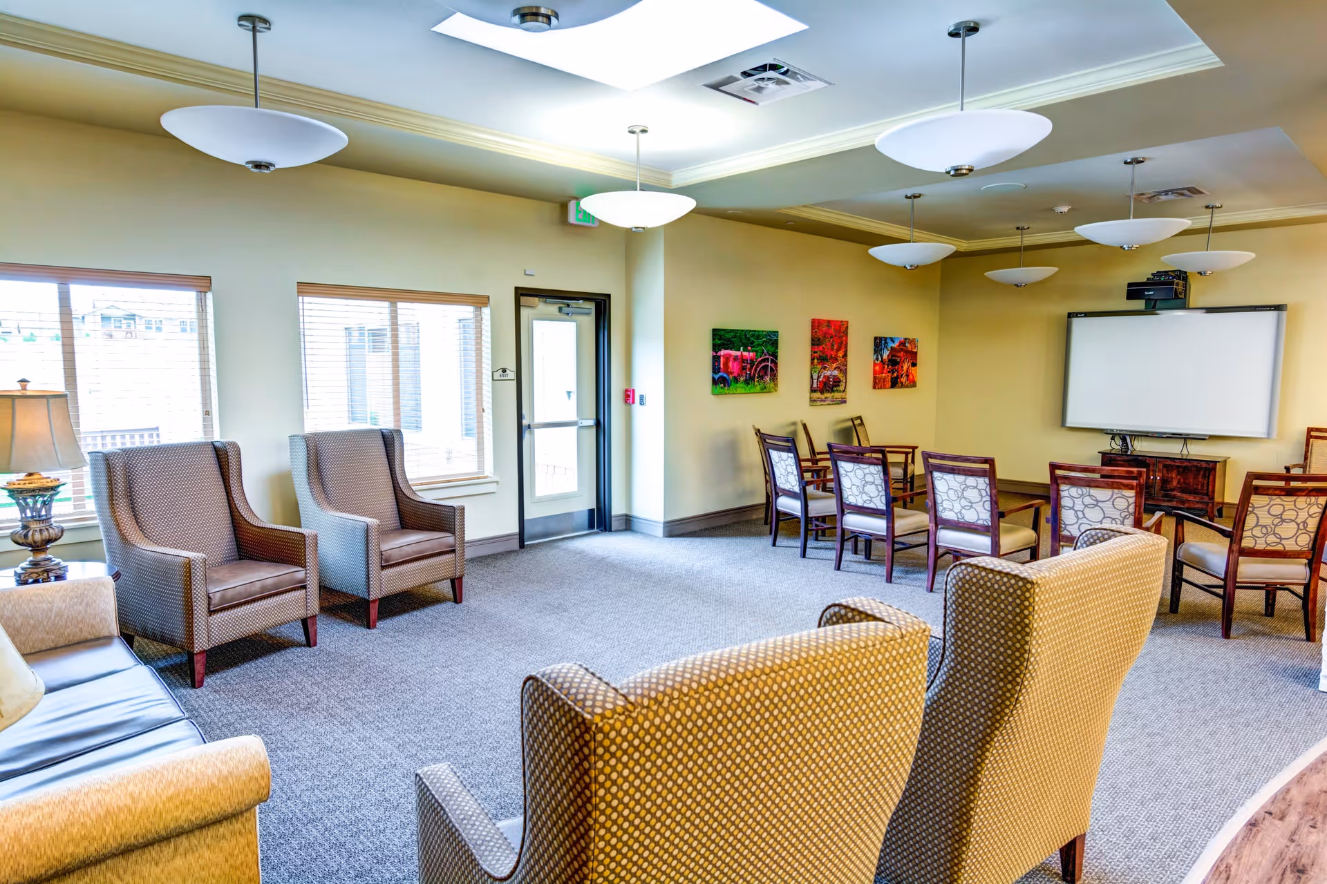 A spacious and well-lit common area with several upholstered armchairs and a sofa arranged around the room. There are large windows letting in natural light, a door leading outside, and a wall-mounted projector screen with chairs arranged in rows facing it. The walls are decorated with colorful artwork, and modern ceiling lights hang from the ceiling.