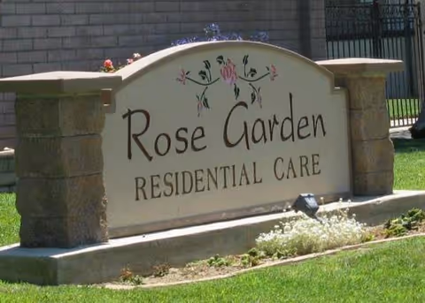 A stone and concrete sign for Rose Garden Residential Care with floral decorations on top, set on a grassy area with a brick wall and metal gate in the background.