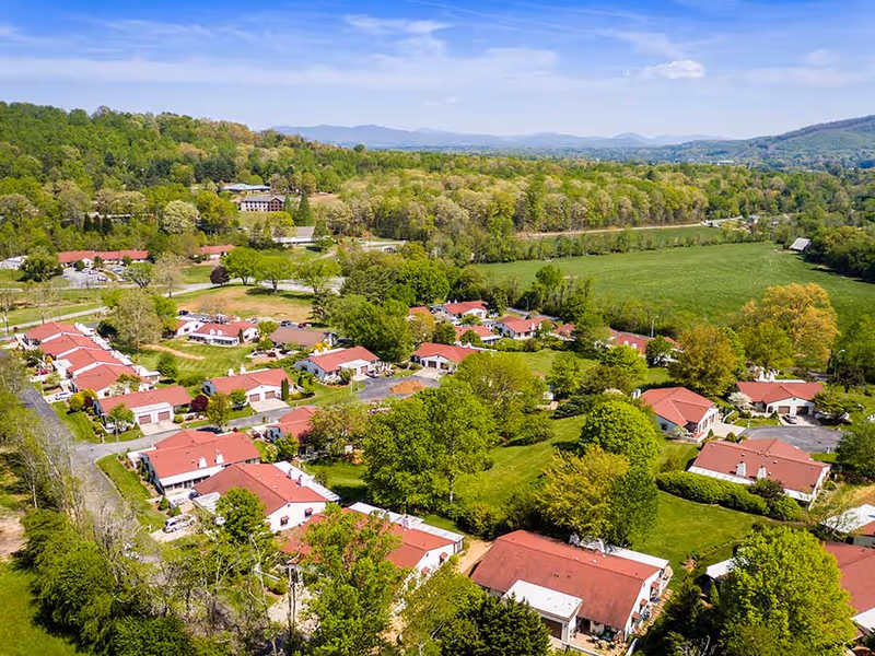 Aerial view of Pisgah Villa at Pisgah Valley Retirement Community showing multiple single-story buildings with red roofs surrounded by green trees and open fields under a blue sky with some clouds.