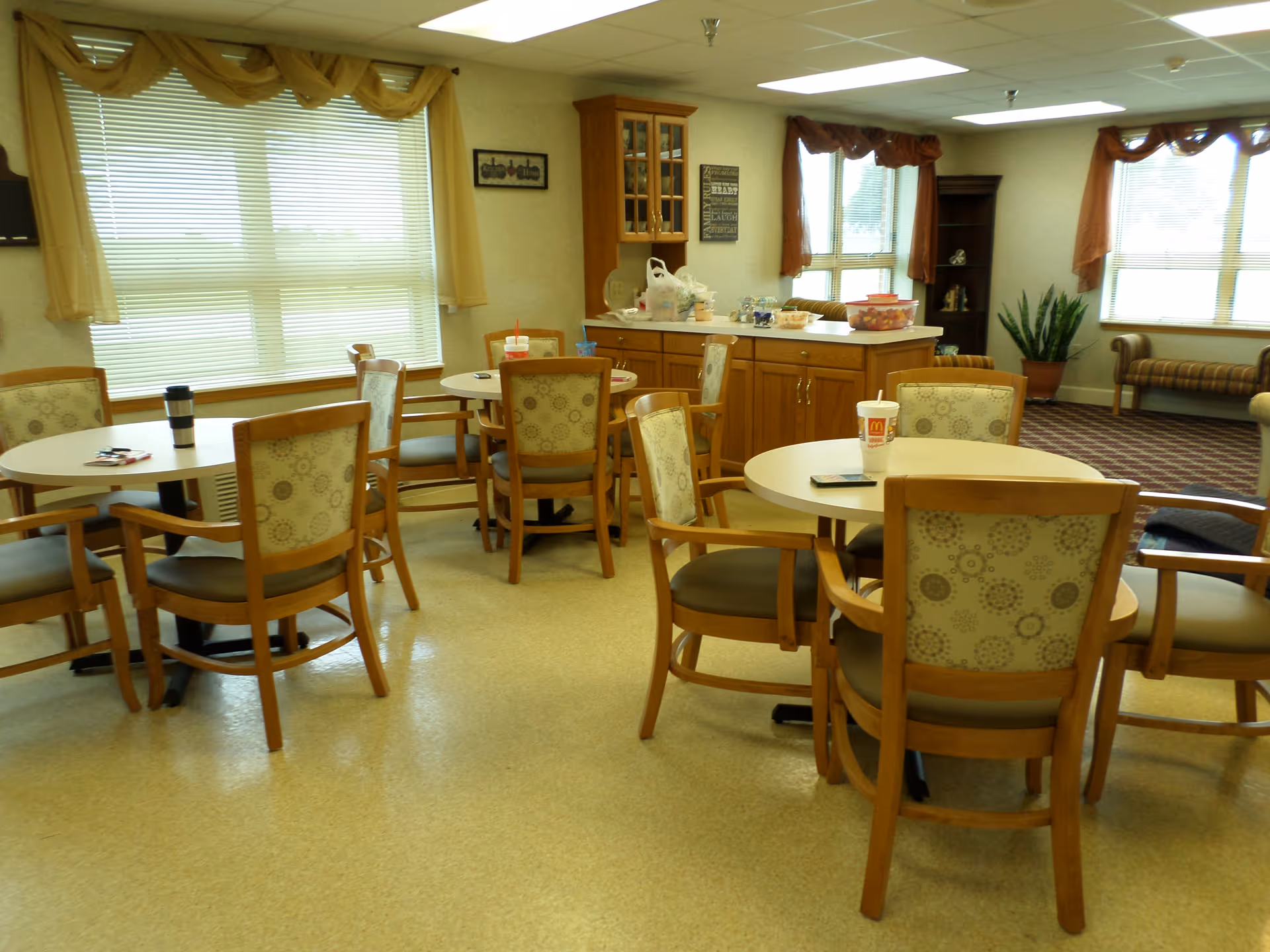 A bright dining area with several round tables and wooden chairs with patterned upholstery. The room has large windows with blinds and valances, a wooden cabinet with glass doors, and a countertop with various items including food containers and drinks. In the background, there is a seating area with a bench and a potted plant near the windows.