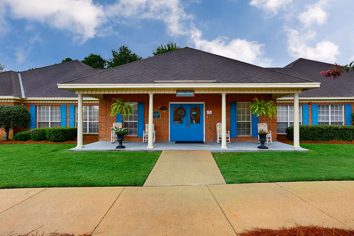 Front exterior view of a single-story brick building with a dark shingled roof and blue double doors labeled Magnolia Cottage. The entrance has a covered porch with white columns, hanging ferns, rocking chairs with floral cushions, and potted plants. There is a concrete walkway leading to the entrance, surrounded by well-maintained green grass and shrubs under a partly cloudy sky.