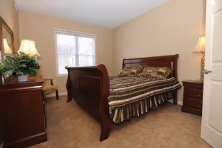 A bedroom with a large wooden sleigh bed covered with a striped comforter and multiple pillows. The room has beige walls and carpeted floor. There is a wooden dresser with a lamp and a plant on it, a chair next to the window, and a nightstand with a lamp beside the bed.