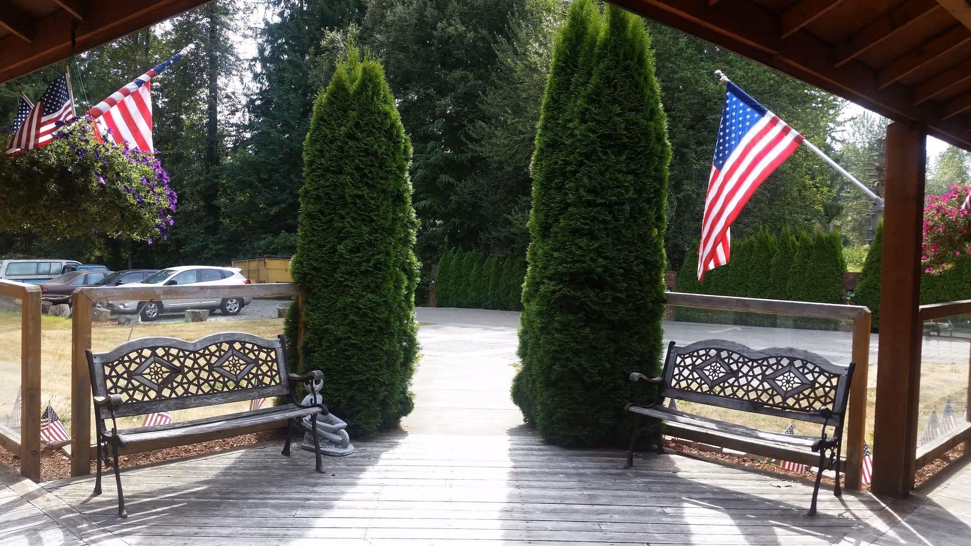 Covered outdoor seating area with two decorative metal benches facing a paved pathway flanked by tall green shrubs. American flags and hanging flower baskets decorate the wooden structure overhead. Several parked cars and trees are visible in the background.