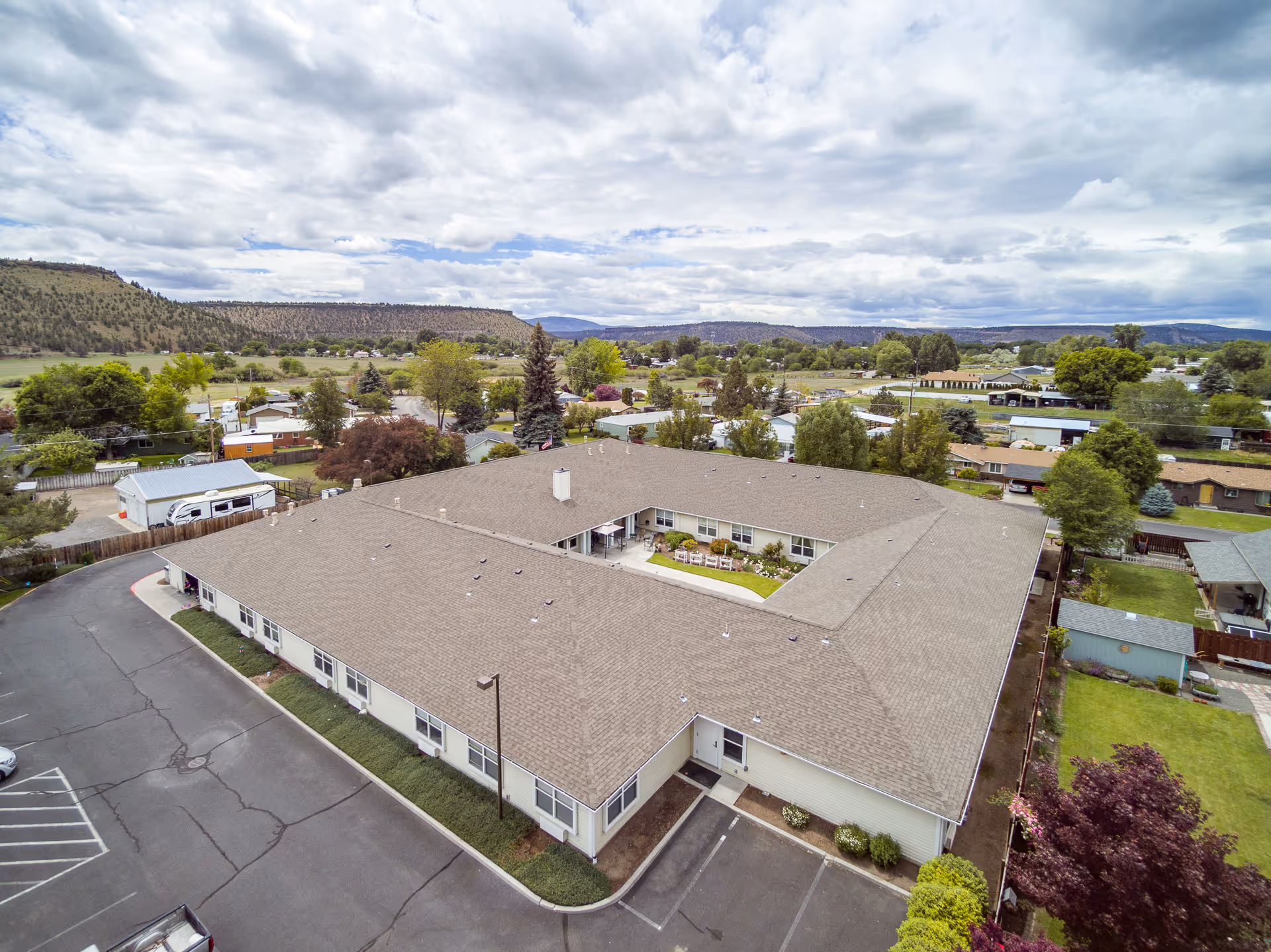 Aerial view of a single-story senior living facility building with a central courtyard, surrounded by parking spaces and greenery, set against a backdrop of hills and cloudy sky.