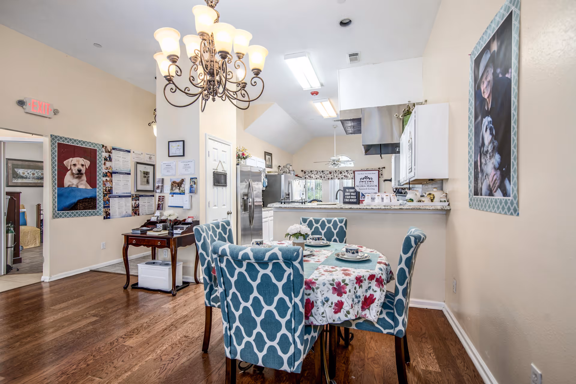 Interior view of a dining area in Acorn Manor Assisted Living featuring a round table with a floral tablecloth and four blue patterned chairs. The room has hardwood floors, a chandelier overhead, and a kitchen area with stainless steel appliances in the background. Walls are decorated with pictures, including one of a dog and another of an elderly person with a dog. There is an exit sign visible on the left side.